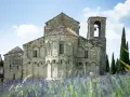 La Pieve di Romena, un'antica chiesa in pietra con campanile, si erge maestosa tra cipressi e un campo fiorito di lavanda sotto un cielo azzurro.