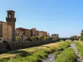 Pescia: cosa vedere? Un fiume serpeggia tra case colorate e un'antica torre con orologio, sotto un cielo azzurro.