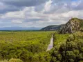 Un fiume serpeggia tra fitti boschi verdi, un paesaggio tipico dei parchi della Toscana, con una torre antica su una collina rocciosa.