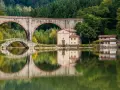 Pieve Fosciana: due ponti, uno antico e uno moderno, si specchiano in un lago calmo, con edifici storici e una foresta sullo sfondo.