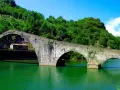 Il Ponte del Diavolo, con i suoi archi in pietra, attraversa un fiume verde smeraldo, circondato da colline lussureggianti e un cielo azzurro.
