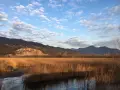 Acque calme del lago di Porta riflettono un cielo azzurro con nuvole bianche, circondato da canneti dorati e montagne all'orizzonte.