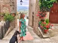 Una donna con un cane passeggia in un vicolo fiorito di Civitella Val di Chiana, con una vista panoramica sullo sfondo.