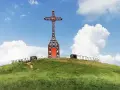 Croce metallica rossa sulla cima di una collina erbosa, con un sentiero che sale. Il **Pratomagno Arezzo** domina il paesaggio sotto un cielo azzurro con nuvole bianche.