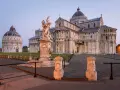 Il **Battistero di Pisa** e il Duomo illuminati dalla luce dell'alba. Una fontana con putti al centro della piazza.
