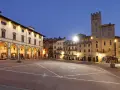 Piazza illuminata di Arezzo al crepuscolo, con portici, negozi e una torre medievale. Qui la vita è bella.