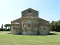 La Basilica di San Piero a Grado, con le sue absidi romaniche e il prato verde circostante, si staglia contro un cielo limpido.
