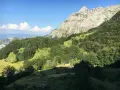 Un piccolo rifugio solitario ai piedi del **Puntato Apuane**, circondato da boschi verdi e montagne imponenti sotto un cielo azzurro.