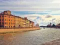 Edifici colorati lungo il fiume Arno a Pisa, con un ponte e nuvole suggestive nel cielo. Un'immagine che cattura la bellezza di Pisa e dintorni.