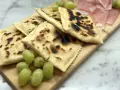 Tortelli alla lastra casentino, dorati e leggermente bruciacchiati, serviti con uva fresca e affettati di prosciutto su un tagliere di legno.