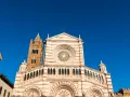 Il Duomo di Grosseto, con la sua facciata a strisce bianche e rosa e il campanile in mattoni, si staglia contro un cielo azzurro intenso.
