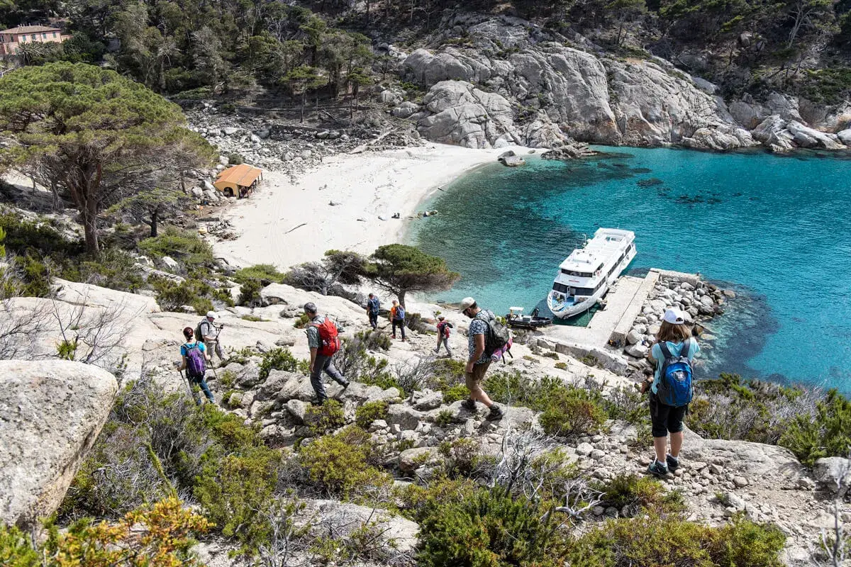 Escursionisti esplorano i sentieri rocciosi dell'isola di Montecristo, con una spiaggia bianca e acque turchesi. Una nave attende nel porto naturale.