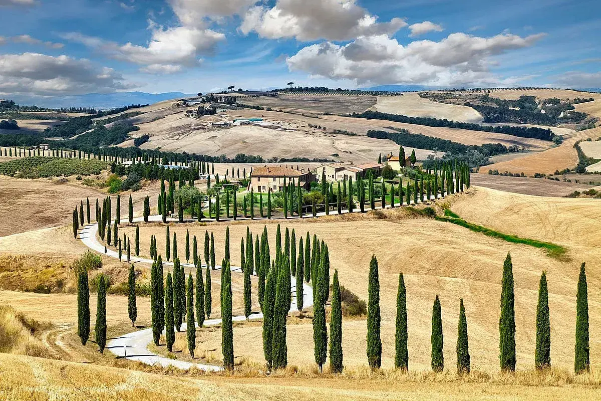 Un casale nel Val d'Orcia, circondato da cipressi e dolci colline dorate sotto un cielo azzurro con nuvole bianche.