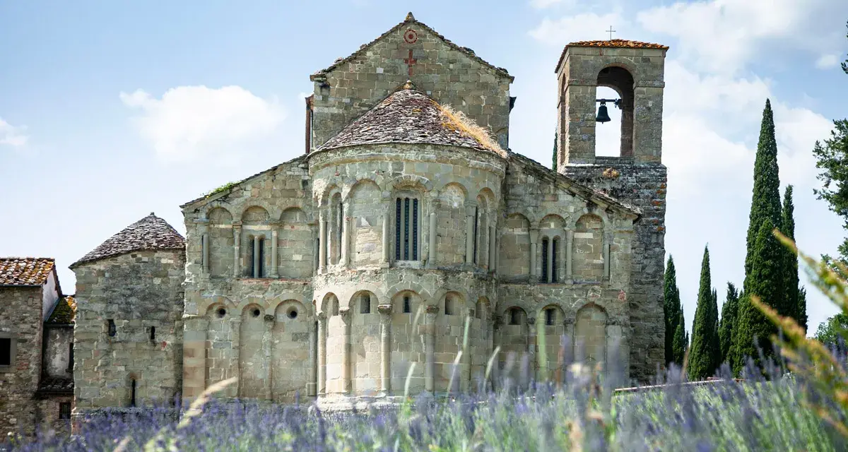 La Pieve di Romena, un'antica chiesa in pietra con campanile, si erge maestosa tra cipressi e un campo fiorito di lavanda sotto un cielo azzurro.
