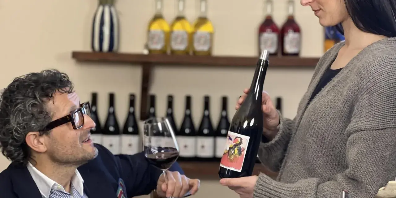 Uomo con occhiali e bicchiere di vino rosso, donna presenta bottiglia. Degustazione vini Arezzo in cantina.