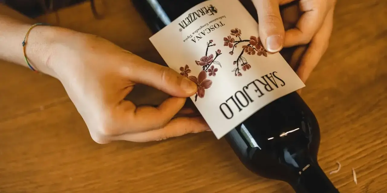 Mani che applicano un'etichetta su una bottiglia di vino. L'etichetta indica "Toscana", la regione di origine del vino Sangiovese.