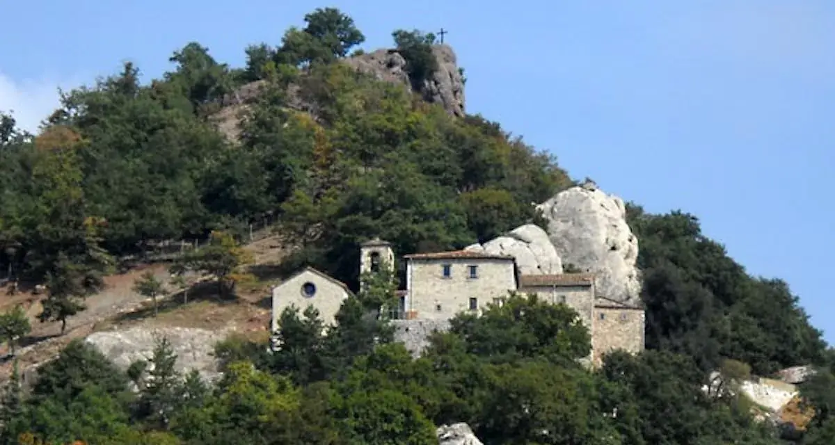 Antico eremo di Cerbaiolo, un complesso di edifici in pietra arroccato su un colle boscoso, con una croce sulla cima di una roccia.