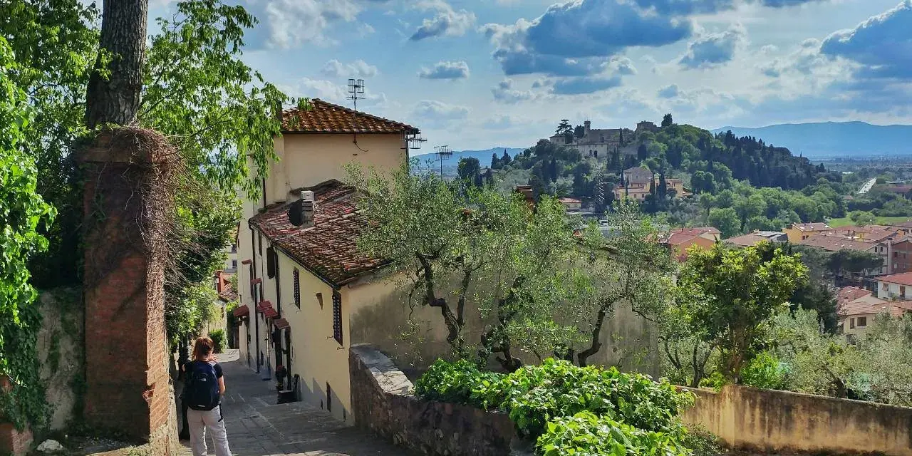 Una persona scende una scalinata in pietra, con case antiche e vegetazione rigogliosa. In lontananza, il **monte Morello** domina il paesaggio collinare.
