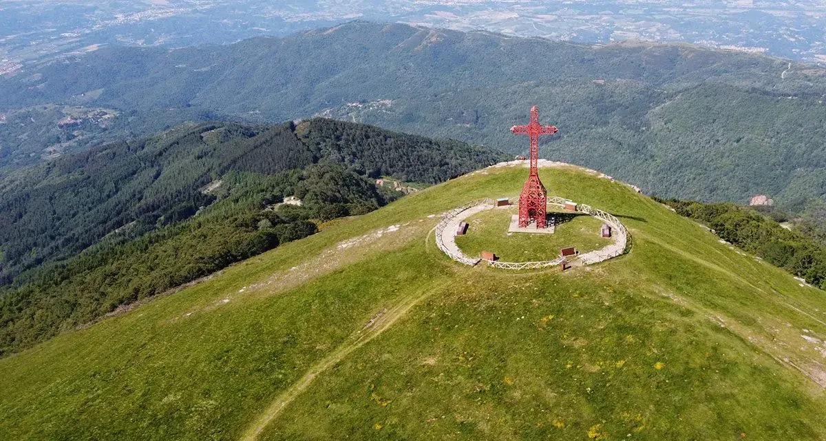La croce del Pratomagno domina un paesaggio verdeggiante, con montagne boscose e una città in lontananza.
