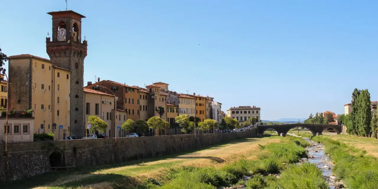 Pescia: cosa vedere? Un fiume serpeggia tra case colorate e un'antica torre con orologio, sotto un cielo azzurro.