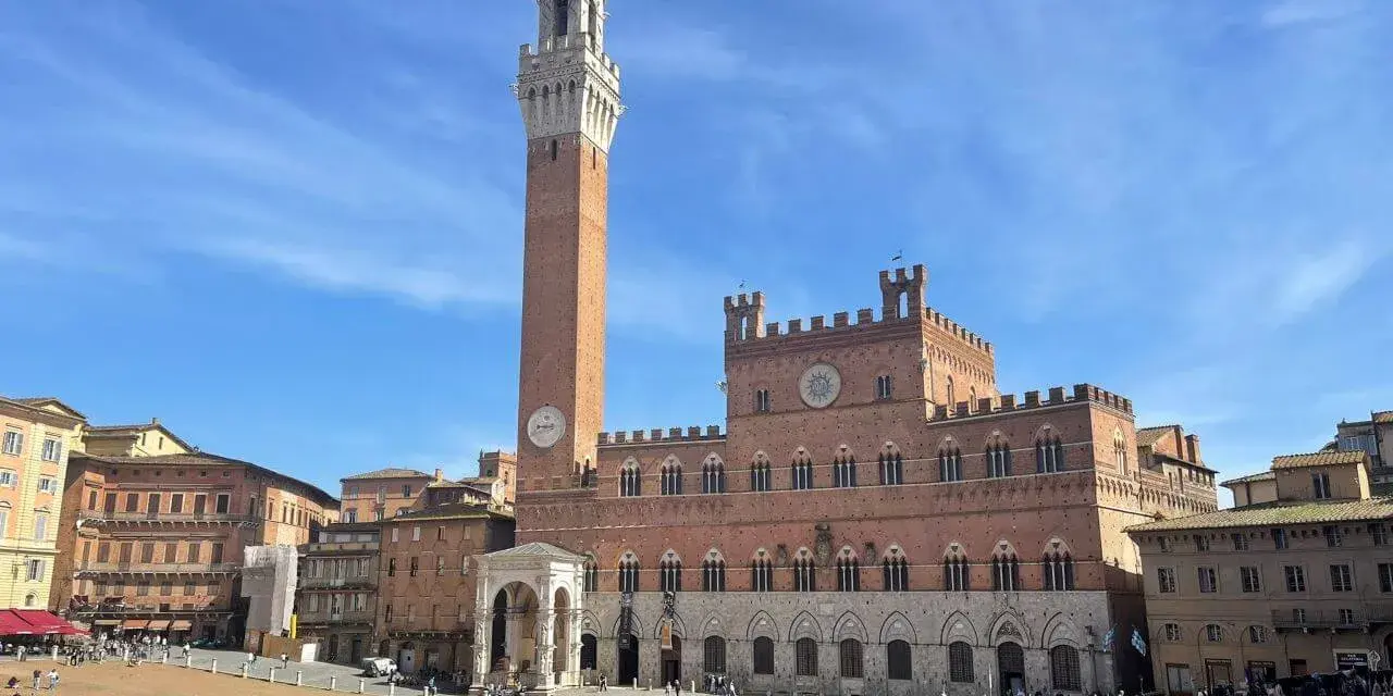 La Piazza del Campo di Siena, con la Torre del Mangia che domina il cielo azzurro, brulica di gente che passeggia e si gode il sole.