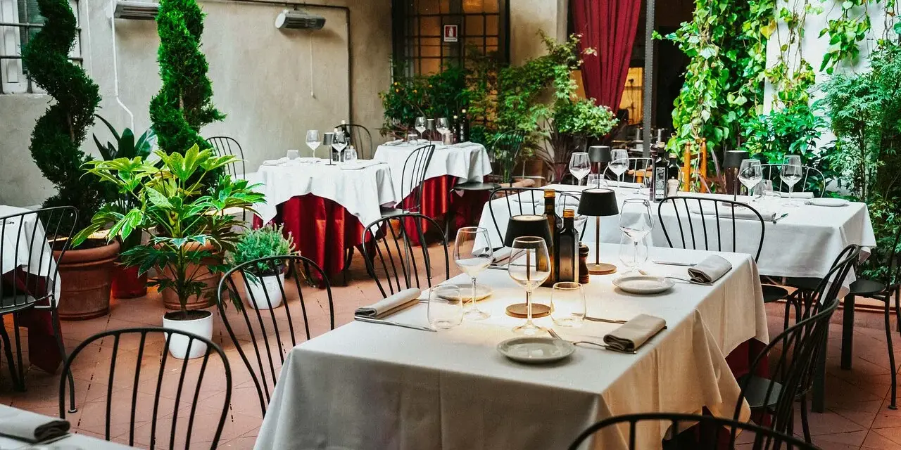 Cortile di un ristorante, tavoli apparecchiati con tovaglie bianche e rosse, piante verdi e sedie nere. Un invito a mangia e bevi santo spirito.