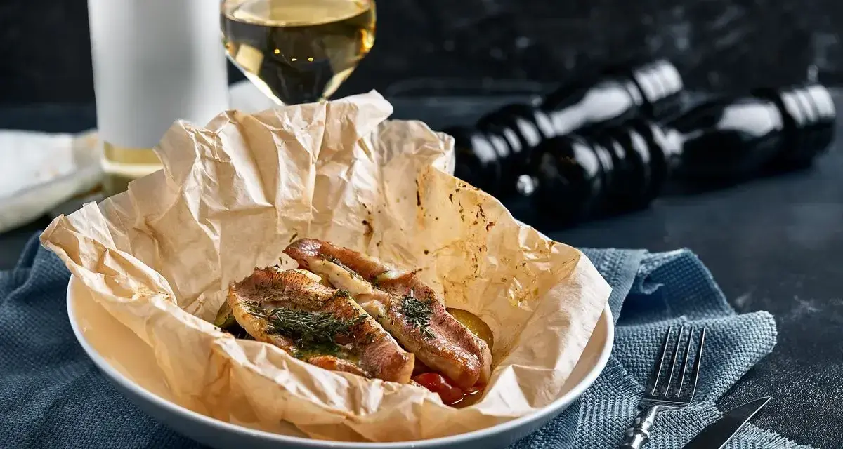 Deliziosa trota puccini cotta al forno, servita su carta da forno con erbe aromatiche. Accanto, un bicchiere di vino bianco e macinapepe.