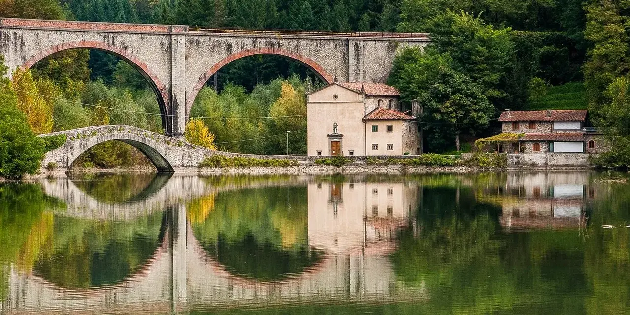 Pieve Fosciana: due ponti, uno antico e uno moderno, si specchiano in un lago calmo, con edifici storici e una foresta sullo sfondo.