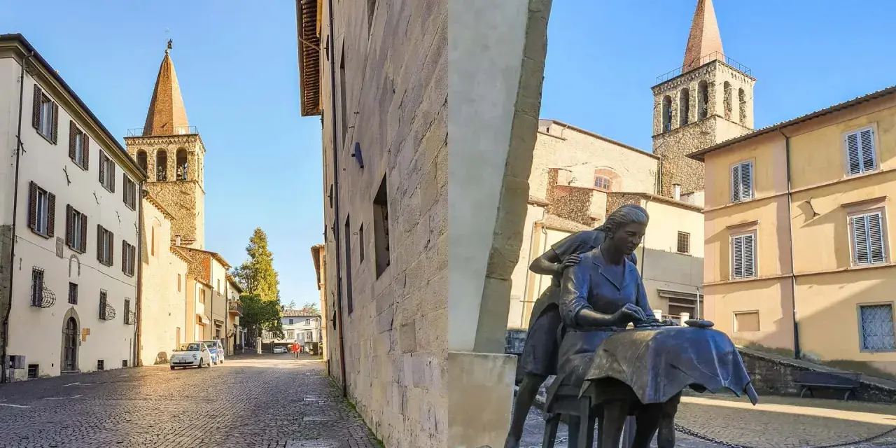 Statua di donne al lavoro, con campanili e palazzi storici sullo sfondo. Un assaggio di cosa vedere a Sansepolcro.