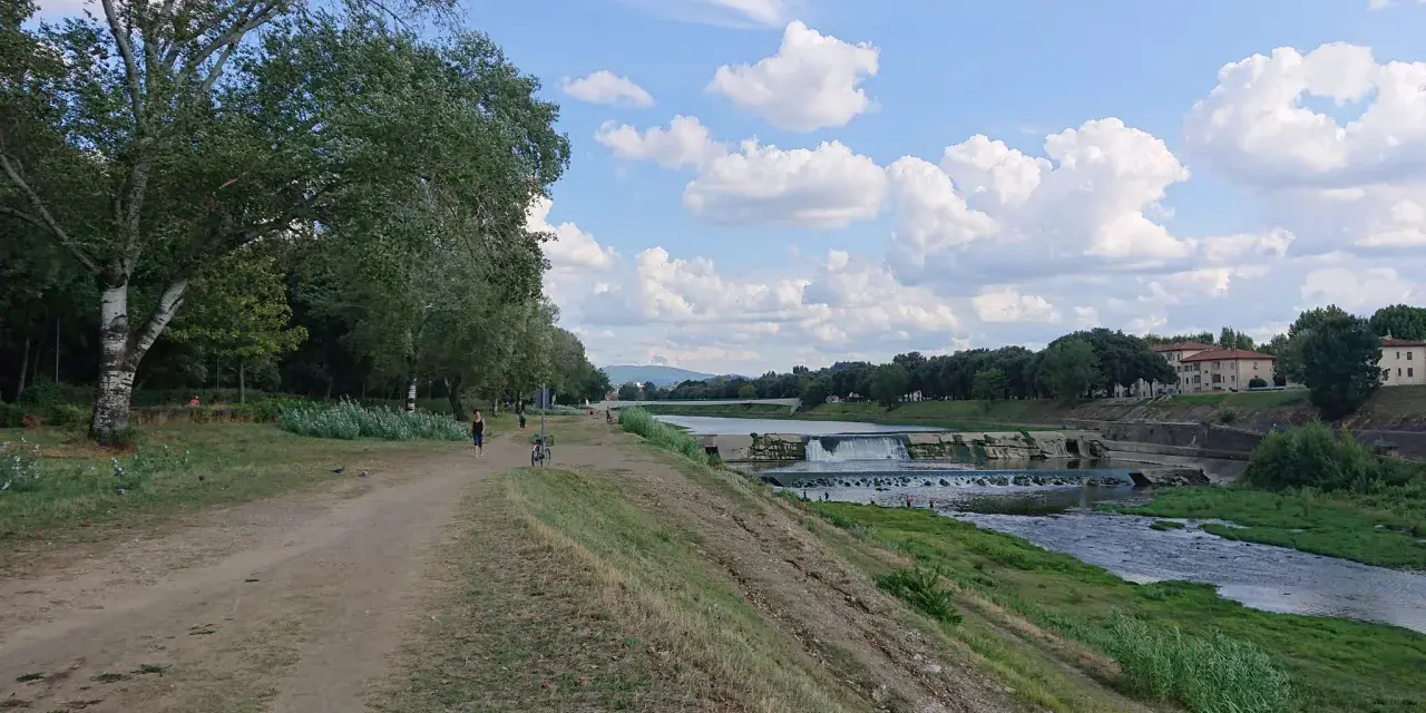 Sentiero lungo il fiume con alberi e il masso della Gonfolina in lontananza. Cielo azzurro con nuvole bianche.