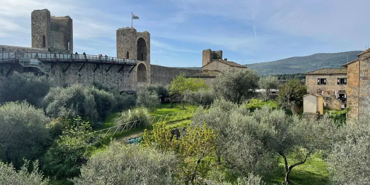 Le mura di Monteriggioni, Siena, dominano un paesaggio toscano con ulivi e colline. Un ponte pedonale attraversa la fortificazione.