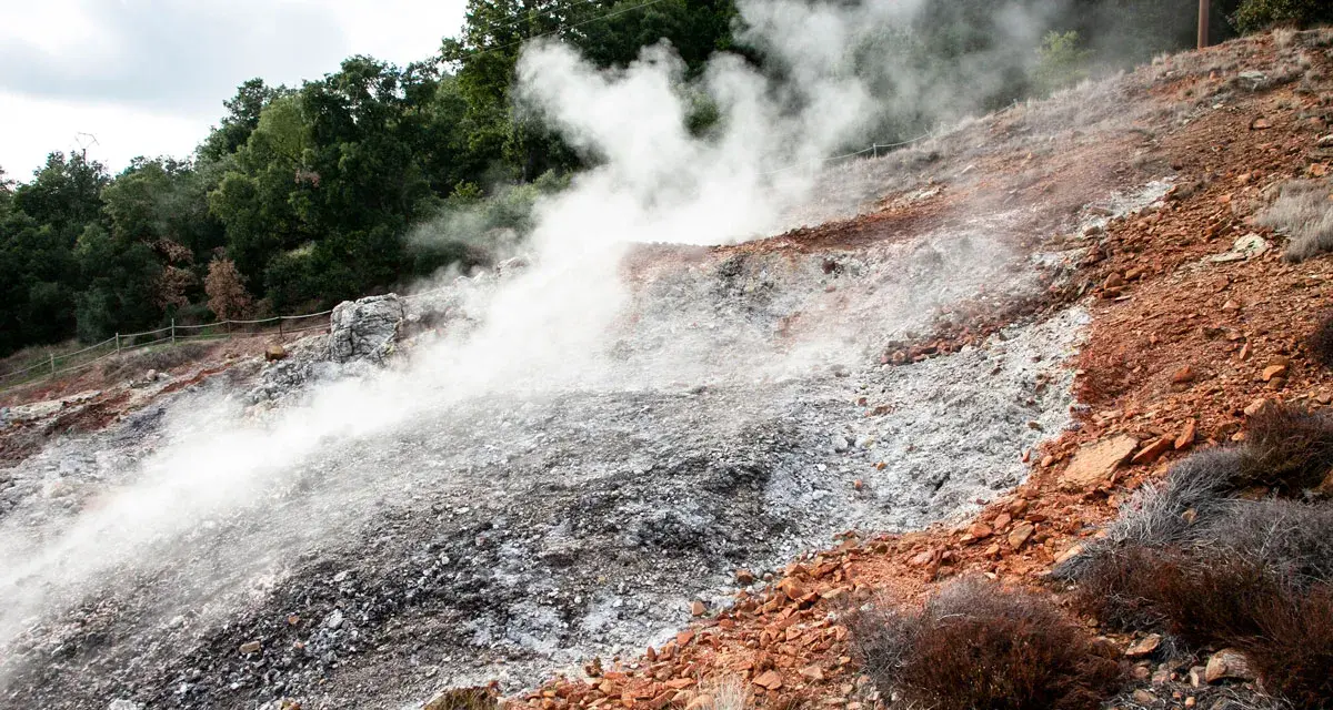 Fumarole attive nel Parco delle Biancane, con vapore che sale da un terreno roccioso e argilloso, circondato da vegetazione e alberi.