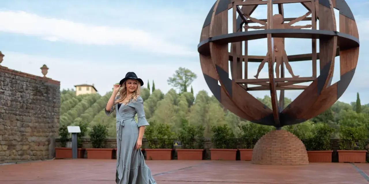 Donna con cappello nero ammira l'Uomo Vitruviano in una sfera di legno, un'opera iconica da vedere a Vinci, con colline verdi sullo sfondo.