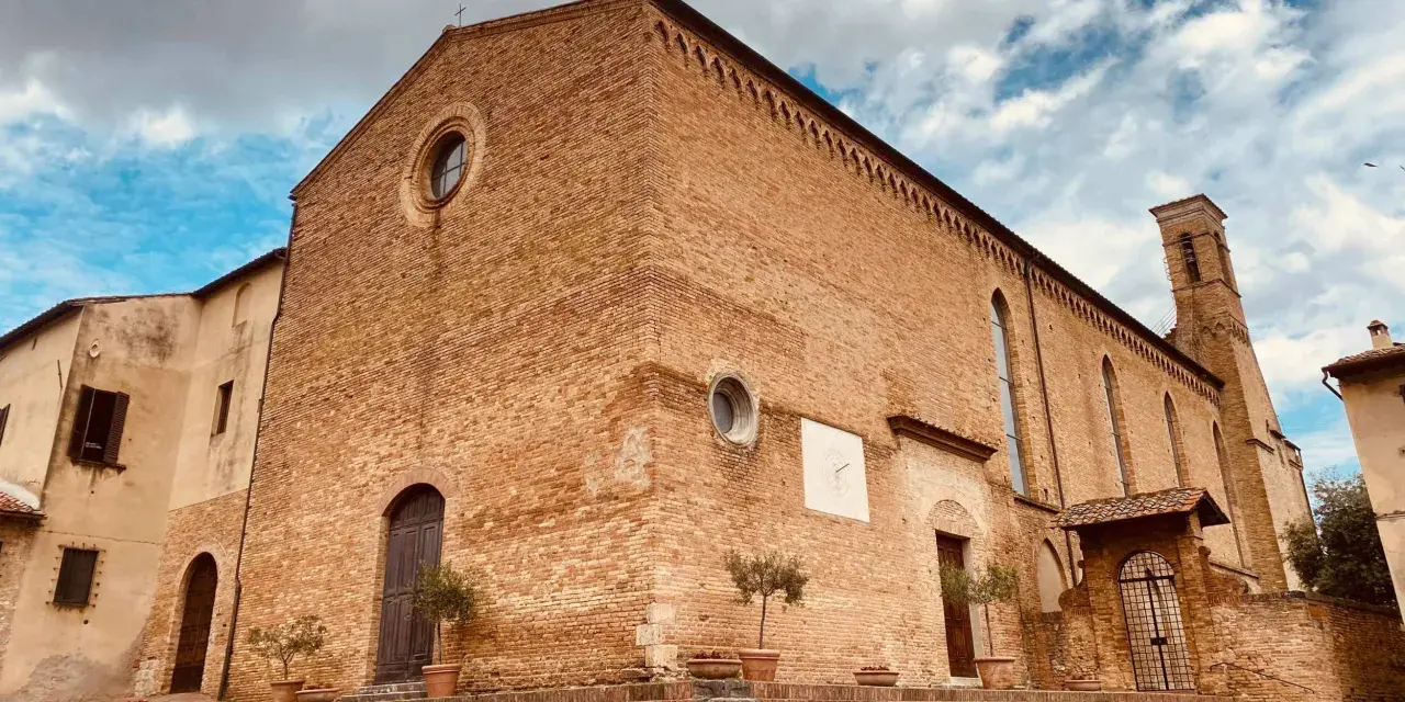 La **chiesa di Sant'Agostino** a San Gimignano, con la sua facciata in mattoni rossi e il campanile svettante, si staglia contro un cielo azzurro e nuvoloso.