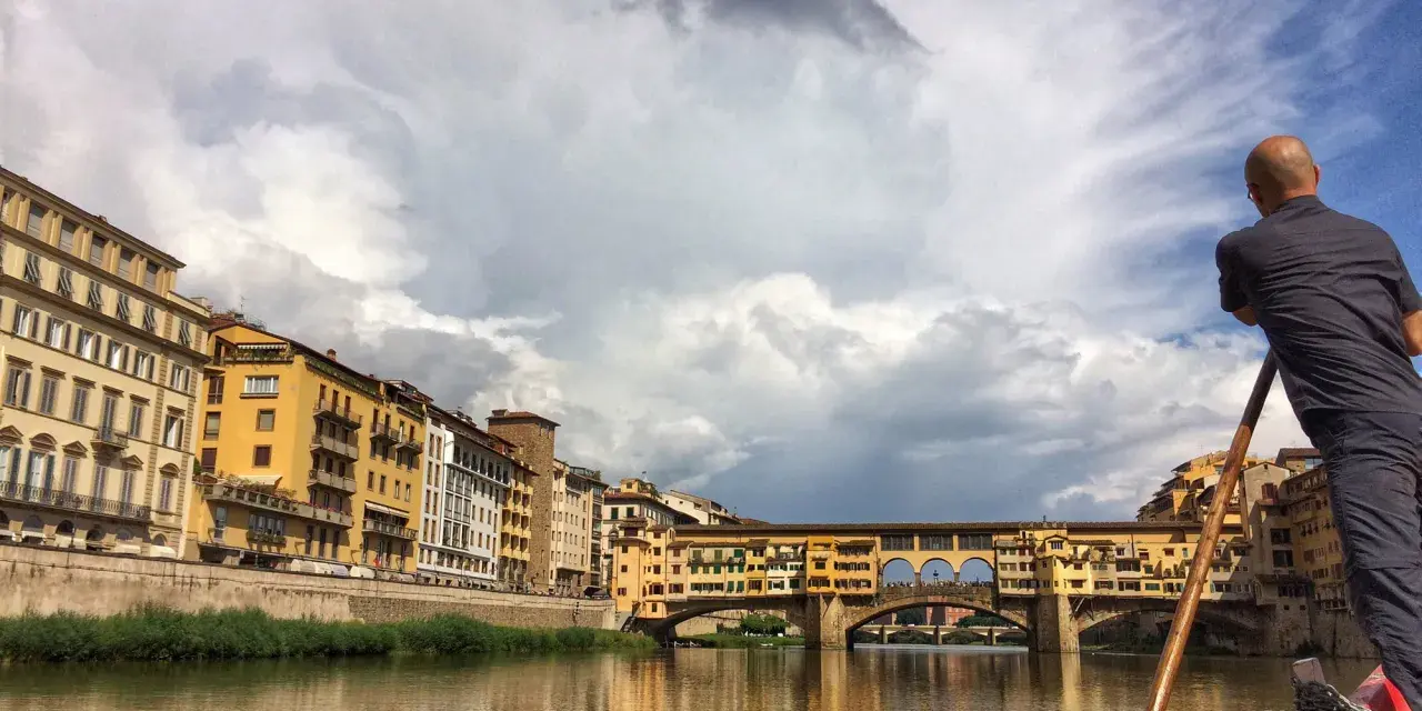 Un uomo su una barca a remi naviga lungo l'Arno, ammirando Ponte Vecchio e gli edifici storici di Firenze.