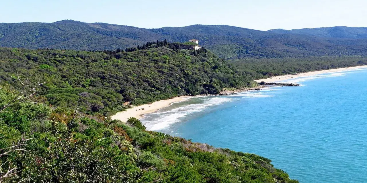 Vista panoramica delle bandite di Scarlino, con una spiaggia dorata bagnata da un mare azzurro e una villa in cima a una collina verde.