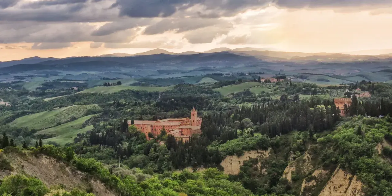 Un imponente complesso monastico, il Monte Oliveto Maggiore, si erge tra dolci colline toscane sotto un cielo drammatico.