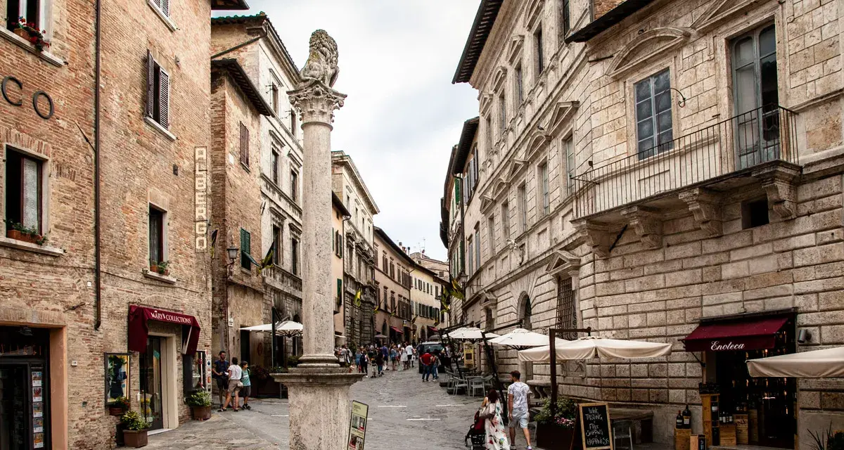 A stone column with a lion statue stands in a cobblestone street lined with historic buildings and shops.