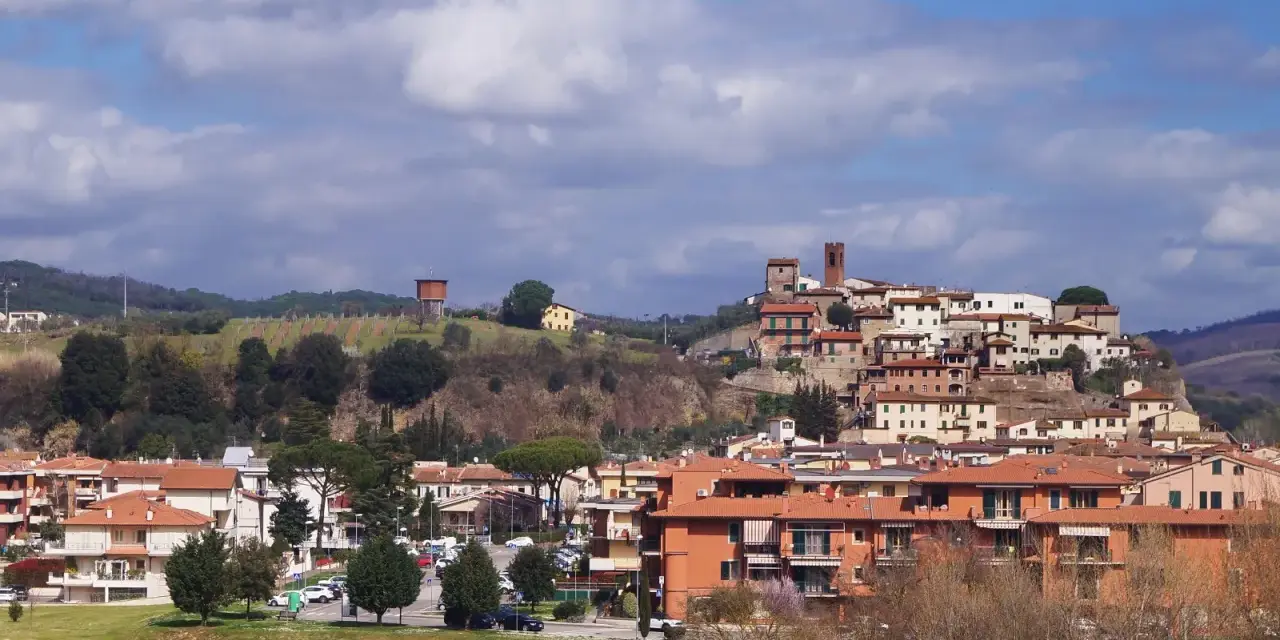 Paesaggio collinare con borgo medievale in cima, case moderne ai piedi. Si intravede un serbatoio d'acqua, vigneti e il **limite** del paese.
