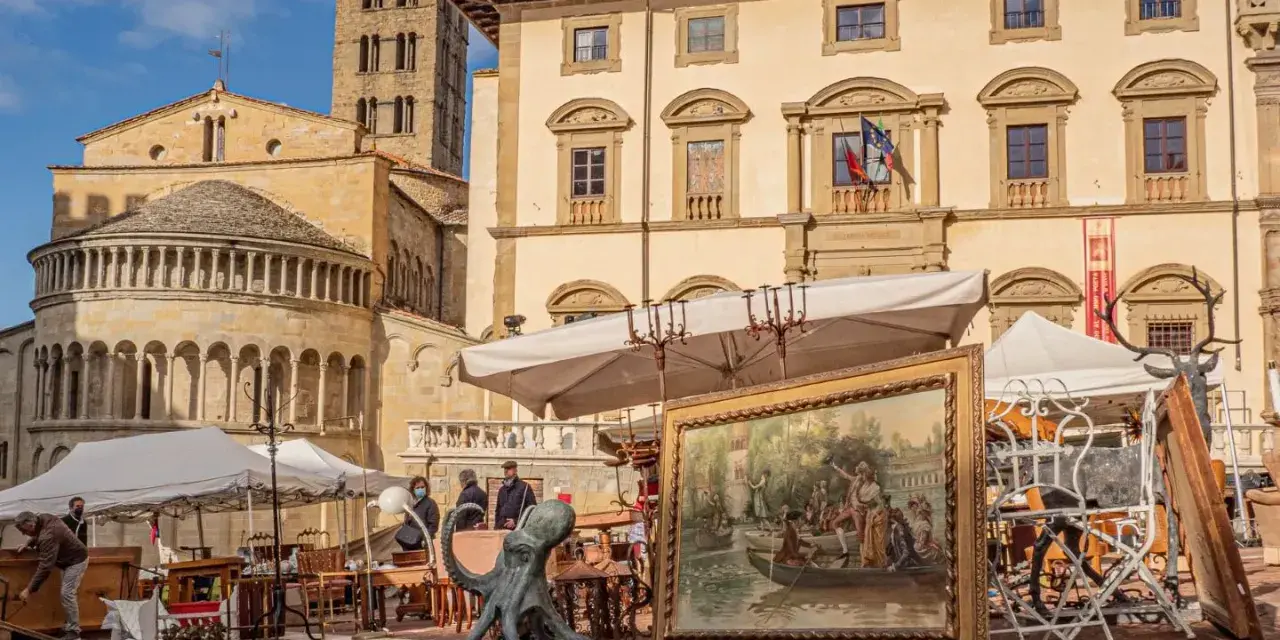 Mercatino dell'antiquariato in piazza grande, con opere d'arte, sculture e mobili antichi esposti sotto il sole.