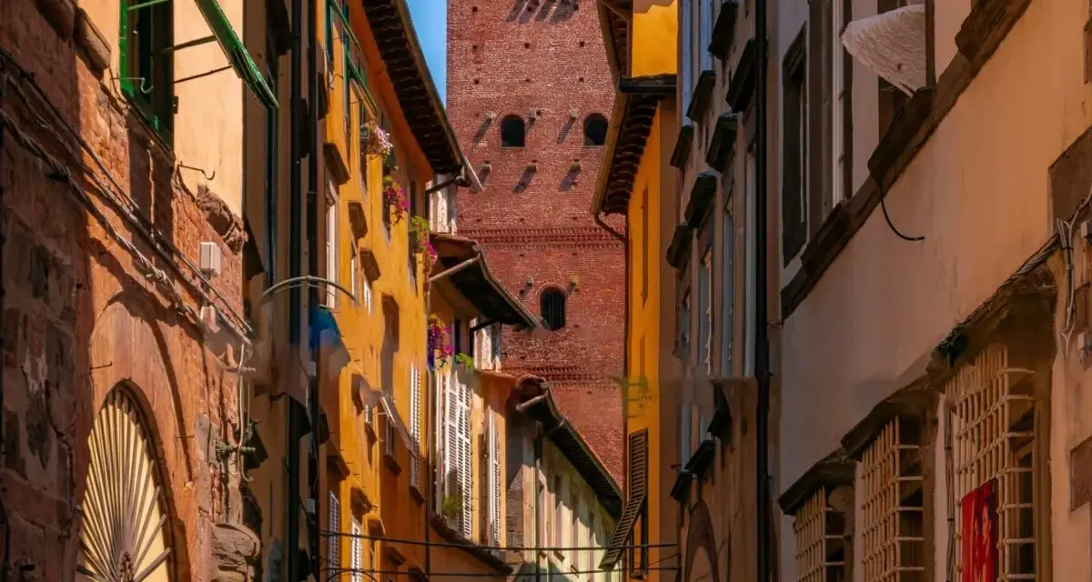 Un vicolo stretto a Lucca con edifici colorati e la torre Guinigi che svetta in lontananza.