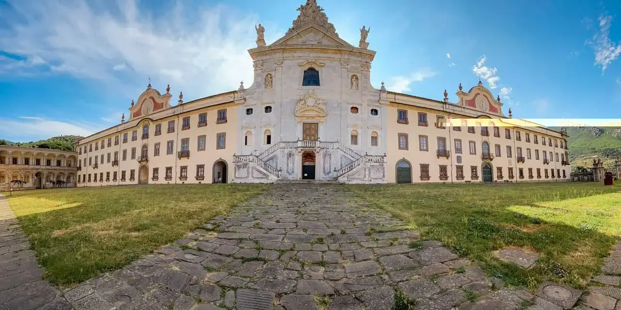 La maestosa Certosa di Calci si erge sotto un cielo azzurro, con un sentiero di pietra che conduce alla sua imponente facciata.