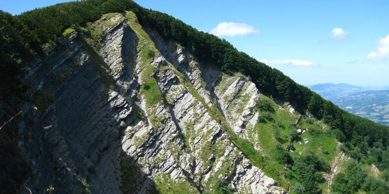 Ripida parete rocciosa con stratificazioni evidenti, coperta da vegetazione verde e alberi. Il cielo è azzurro con qualche nuvola.