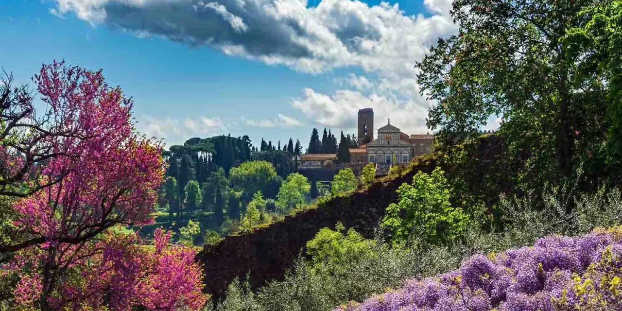 Giardini fioriti con glicini viola e alberi rosa, sullo sfondo un edificio storico con torre: curiosità da vedere a Firenze.