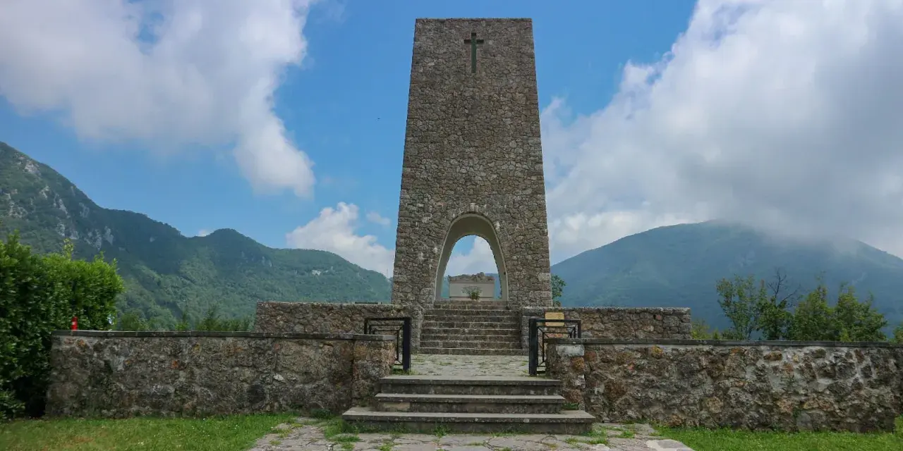 Monumento a Sant'Anna di Stazzema, cosa vedere: un imponente torrione in pietra con arco, che custodisce una tomba, circondato da mura e montagne.