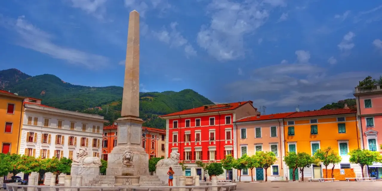 Piazza con obelisco e leoni, edifici colorati e montagne sullo sfondo. Un'idea di cosa vedere a Marina di Massa.
