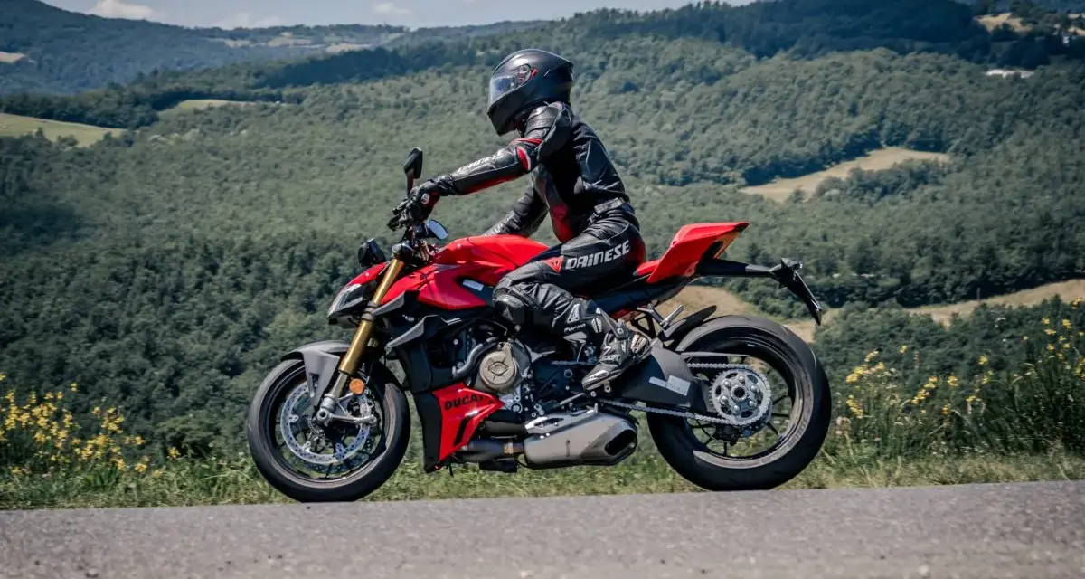 Motociclista in tuta rossa e nera sfreccia su una Ducati rossa lungo il passo del giogo, tra colline verdi e cielo azzurro.