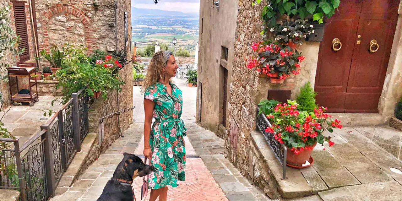 Una donna con un cane passeggia in un vicolo fiorito di Civitella Val di Chiana, con una vista panoramica sullo sfondo.