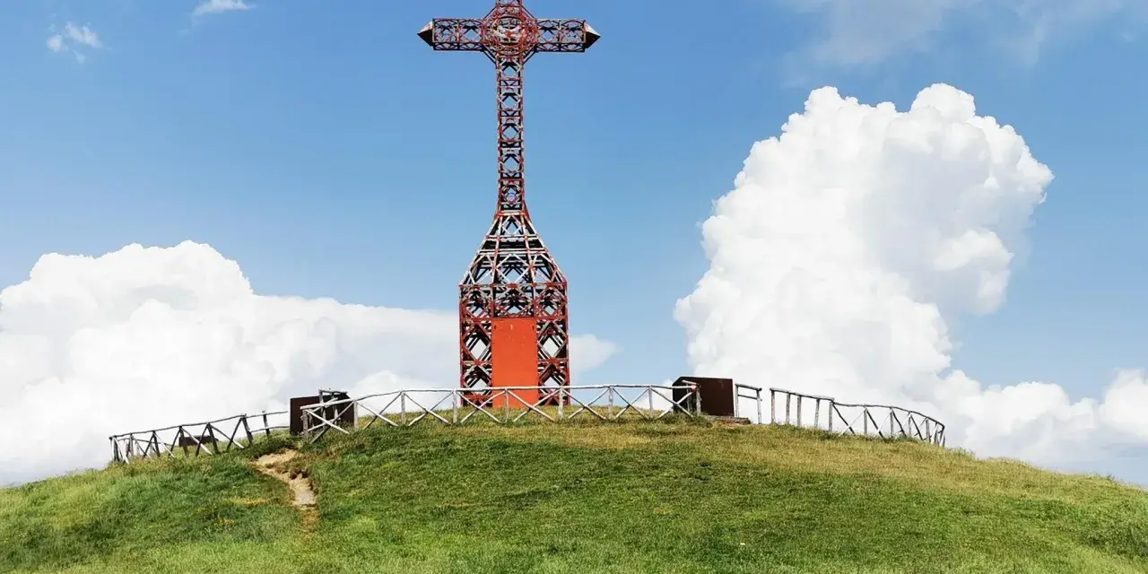 Croce metallica rossa sulla cima di una collina erbosa, con un sentiero che sale. Il **Pratomagno Arezzo** domina il paesaggio sotto un cielo azzurro con nuvole bianche.