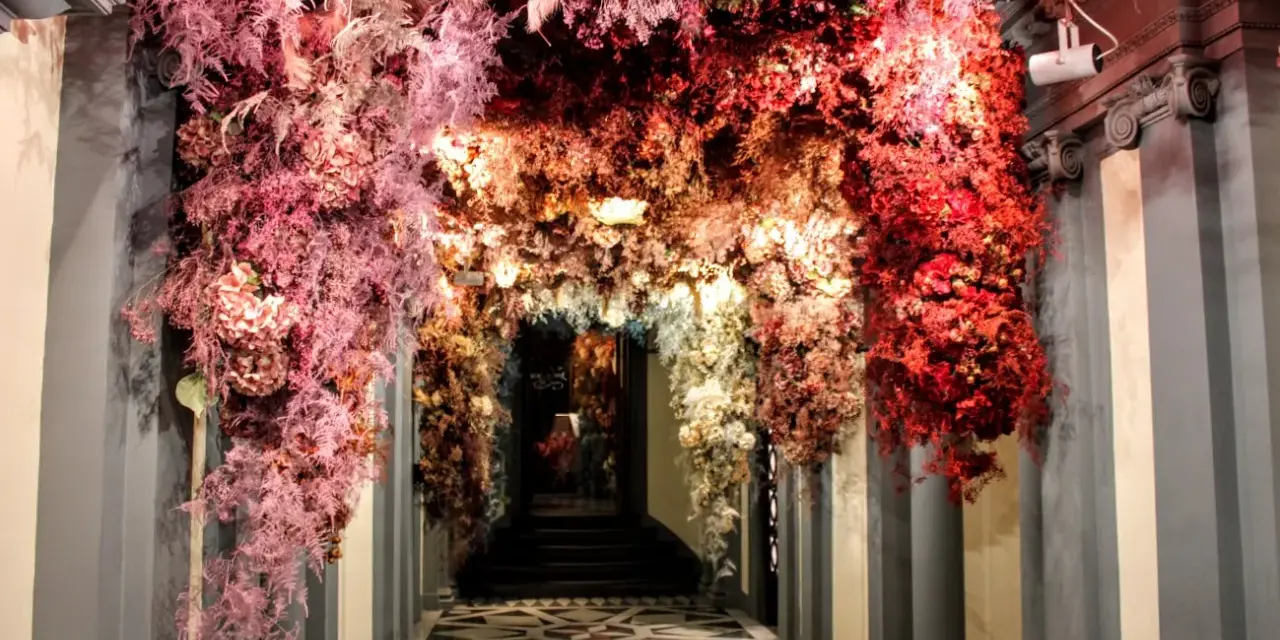 Ingresso fiorito, un arco di fiori rossi e rosa che ricorda l'antica farmacia di Firenze.